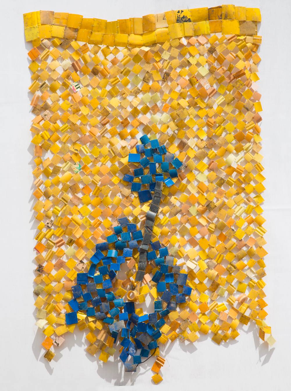 Photograph of a sculpture hanging on a wall. Sculpture is made of small pieces of yellow plastic pieced together like a quilt.